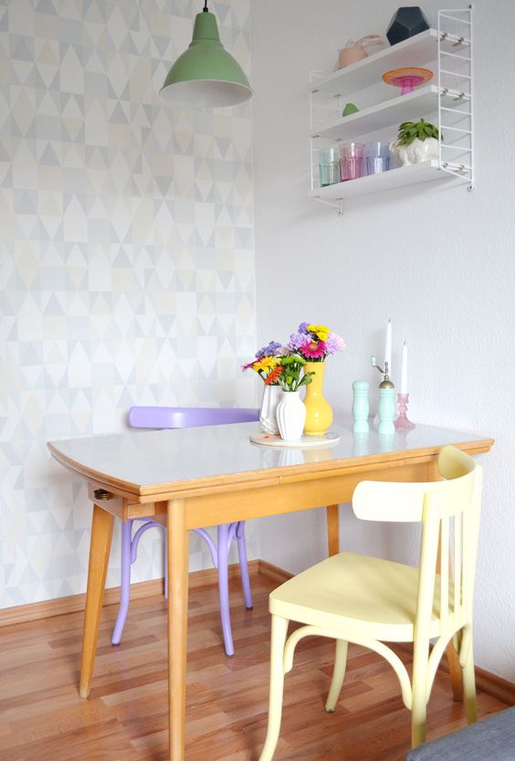 a small dining space with a geometric accent wall, a small table and pastel chairs, a storage unit and a green pendant lamp