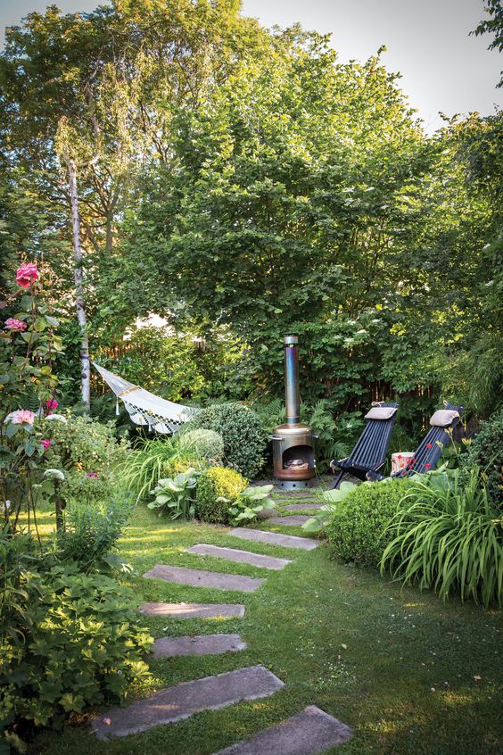 a small garden with a green lawn, greenery, blooms, shrubs and trees, a hearth, some loungers and a hammock