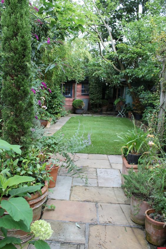 a small modern garden with a green lawn, trees, shrubs, potted greenery and blooms, a tiled pathway is welcoming and cool