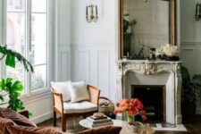 a sophisticated French chic living room with a French fireplace, a large mirror in an ornated frame, a neutral chair, a rust-colored sofa with pillows and potted plants