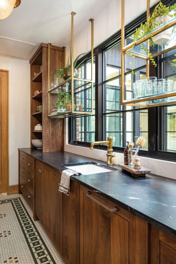 a stained kitchen with black stone countertops and brass suspended shelves over the windows is a cool and elegant space