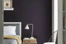 a stylish moody bedroom with a deep purple accent wall, grey and white furniture, a small nightstand with a table lamp and ledges with artworks