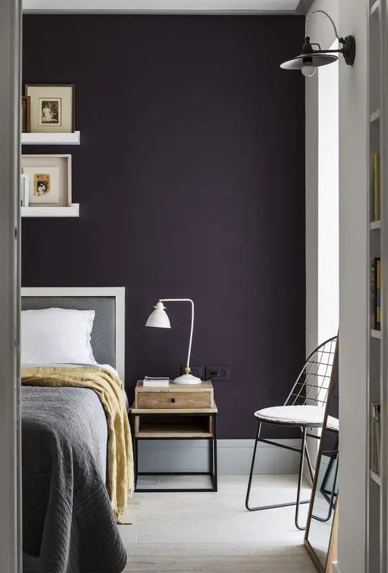 a stylish moody bedroom with a deep purple accent wall, grey and white furniture, a small nightstand with a table lamp and ledges with artworks