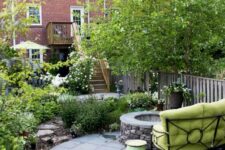 a stylish townhouse garden with a stone platform, a fire pit, a green sofa, planted flowers and greenery