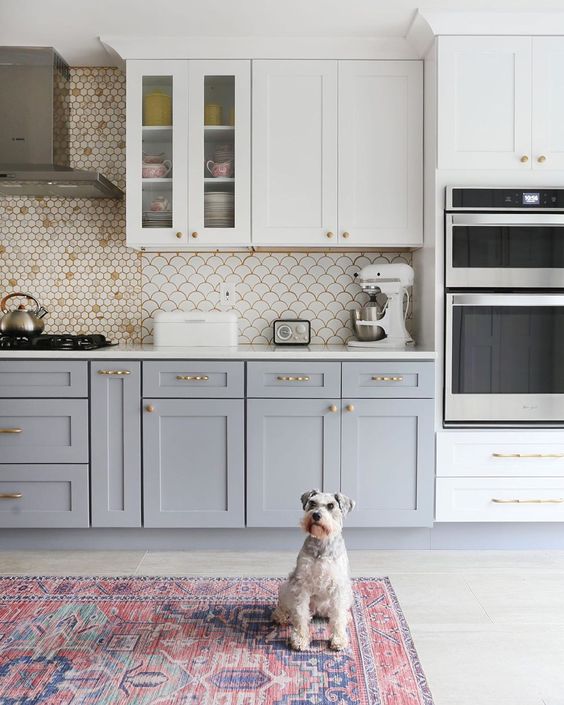 a two-tone kitchen with shaker cabinets, a white scallop backsplash, brass handles and knobs plus a bold rug