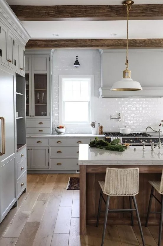 a vintage kitchen with dove grey cabinets, white tile backsplash, white lamps and wooden beams plus a kitchen island