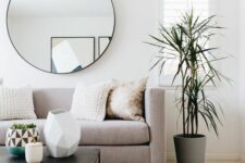 an airy and serene living room with a grey sofa, a black coffee table and vases, some pillows and poufs plus a round mirror