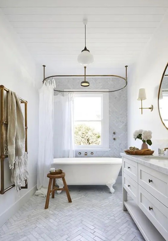 an airy bathroom with a double-hung window, a large vanity, an oval clawfoot bathtub and a marble herringbone tile floor