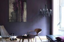 an eclectic living room with a black leather daybed, a couple of wooden chairs and a table, a black chandelier and a deep violet accent wall