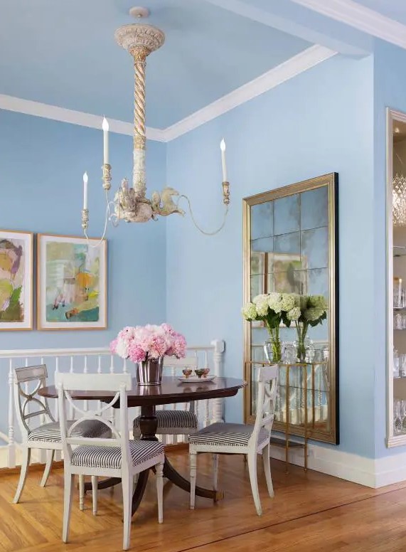 an eclectic pastel blue dining room with a vintage mirror, a stained table and white chairs, a chandelier suspended on rope