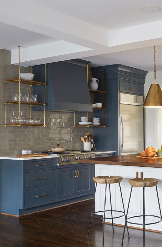 an elegant blue kitchen with a glossy grey tile backsplash, suspended brass shelves, brass pendant lamps and stools
