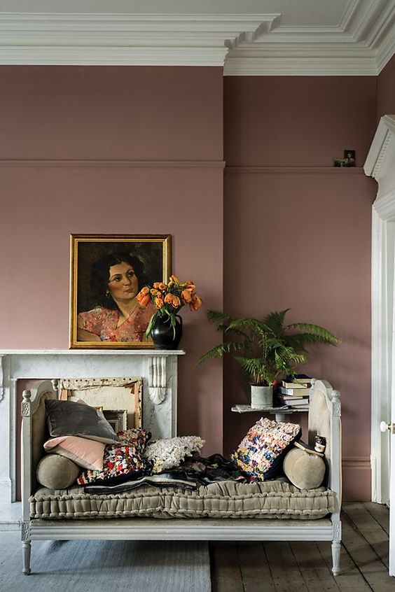 an elegant mauve living room with a firpelace, a vintage daybed with printed textiles, potted plants and artwork