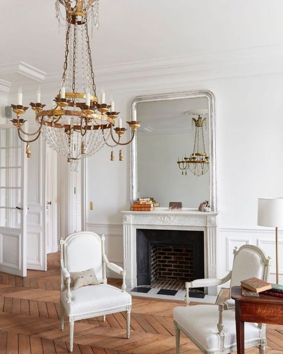 an elegant neutral space wiht a French fireplace, refined vintage chairs, a heavy crystal chandelier and a stained desk