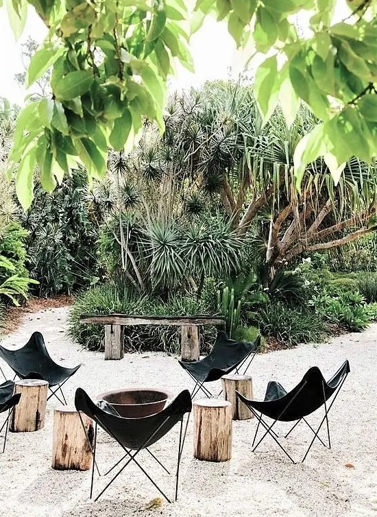 an outdoor fire pit space with a metal fire bowl, black leather butterfly chairs and tree stumps plus greenery around