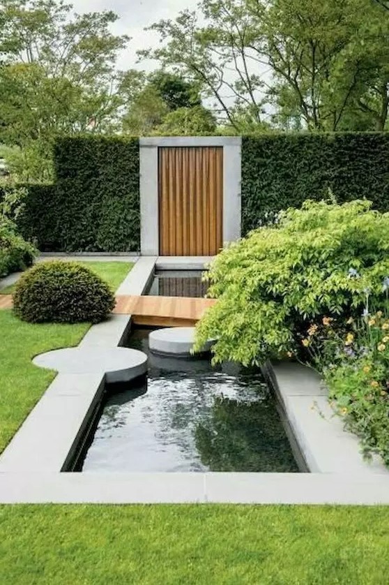 a modern garden with lots of greenery and bushes, with a water feature, stone pavements and a pathway over the water feature