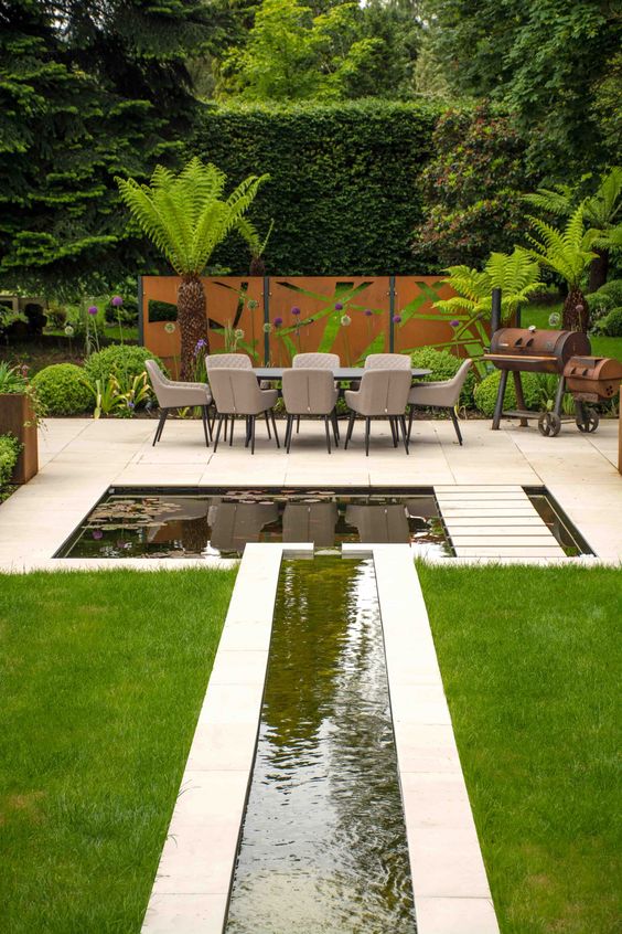 a modern outdoor tropical space with a lawn, greenery and trees, a pond and a waterfall plus a tile deck is all cool
