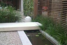 a stylish outdoor space with a modern pond with water plants, gravel pathways, a couple of large stones and greenery around