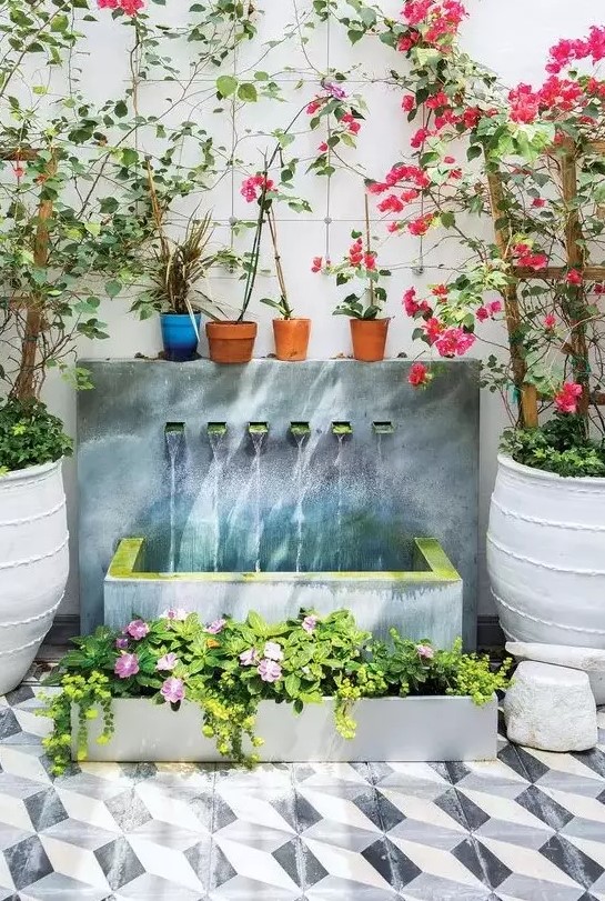a beautiful modern waterfall of a wall with multiple faucets, a concrete tub and a matching planter with blooms next to it