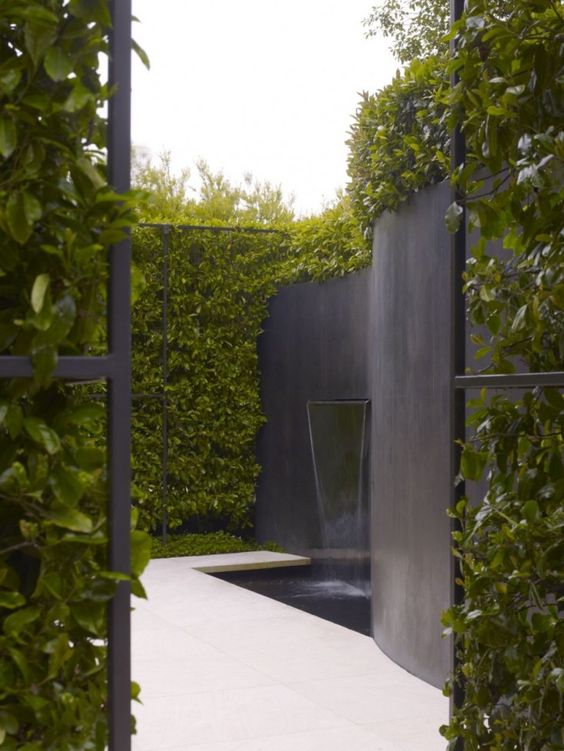 a minimalist outdoor space with greenery walls, a black wall with a waterfall flowing into a water feature and a white tile deck