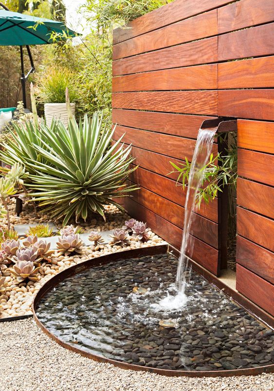a modern outdoor fountain coming of a stained wooden wall into a bowl with pebbles, with succulents and large agaves