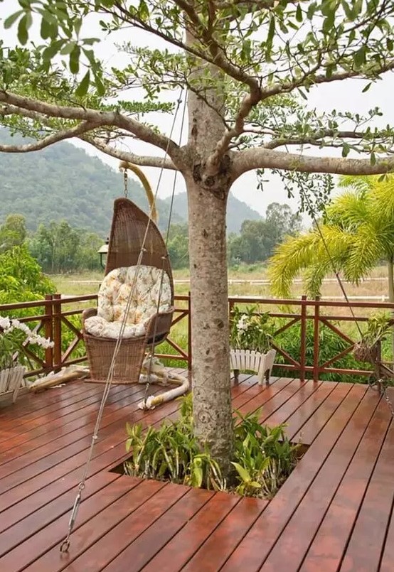 a deck built around a tree, with some greenery surrounding the tree, a wicker pendant chair and some potted blooms