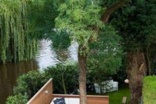 36 a living tree with a deck around it, with white seating furniture and a river behind the deck compose a cool space