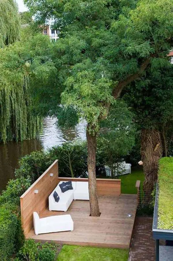 a living tree with a deck around it, with white seating furniture and a river behind the deck compose a cool space