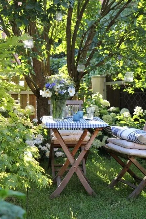 a lawn around the tree used as a little dining space, with stained folding furniture, cushions and pillows, hanging candle lanterns and blooms