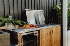a Scandinavian bbq area with a wooden deck and a back wall, a wooden cabinet with a grill and a fridge, some greenery around