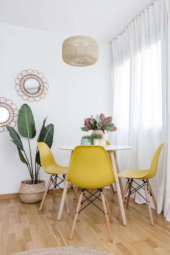 a bold boho dining space with a round table, yellow Eames chairs, a potted plant, a woven pendant lamp and some mirrors