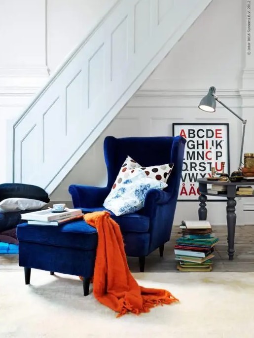 a bold reading nook with a navy Strandmon chair and an ottoman, a a grey table, book stacks and a table lamp is cool