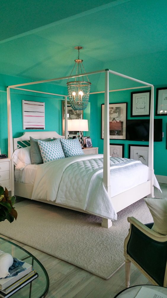 a bold turquoise bedroom with a white canopy bed and neutral bedding, a large gallery wall and a crystal chandelier