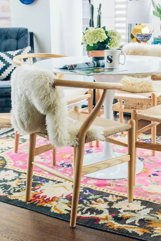 a bright dining room with a round table, light-stained wishbone chairs, a colorful rug and some faux fur
