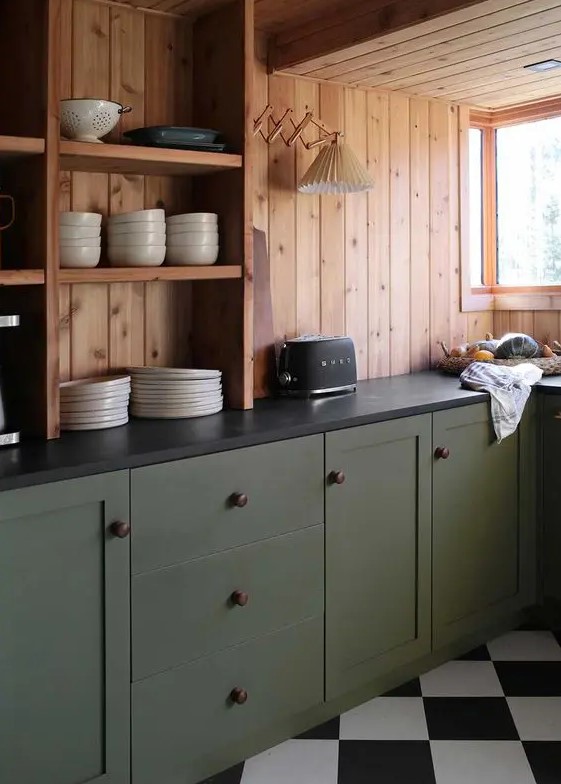 a cabin kitchen with stained wood walls, olive green lower cabinets, black stone countertops and a sconce