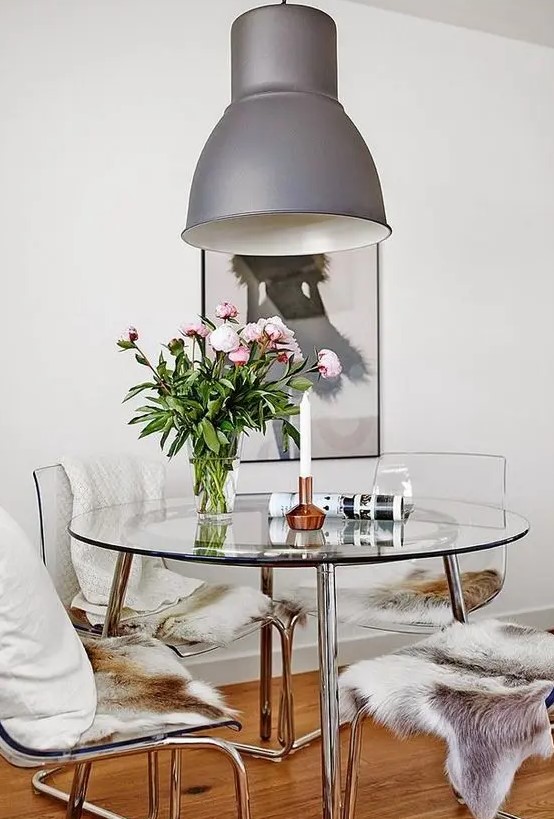 a catchy modern dining space with a round glass table, ghost chairs with faux fur and pillows, a grey pendant lamp