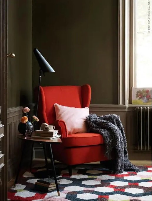 a catchy space with taupe walls and paneling, a red Strandmon chair, a printed rug, a side table and a black lamp is bold and cool