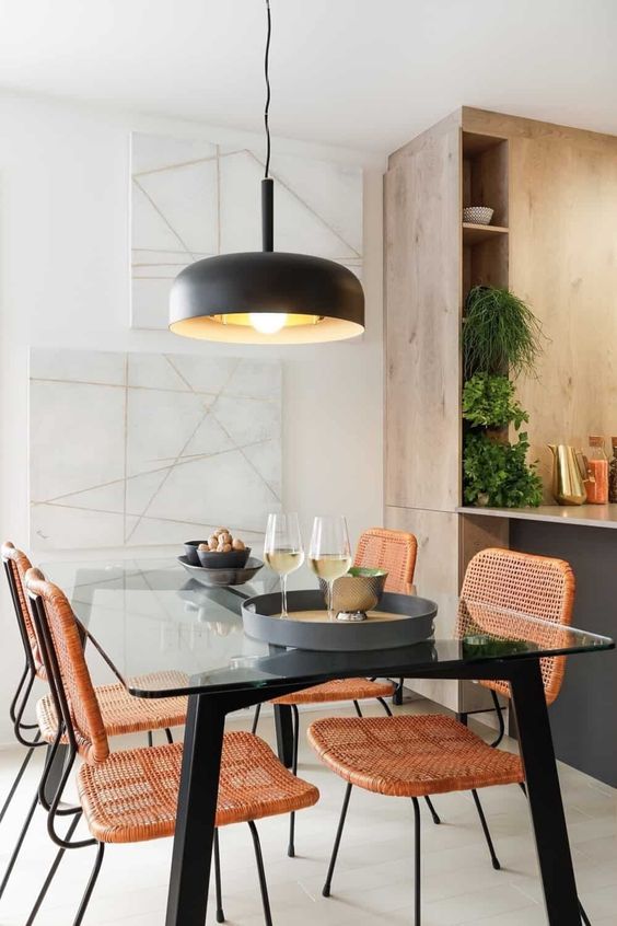 a chic modern dining space with a glass top dining table with black legs, orange cane chairs and a black pendant lamp