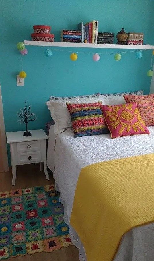 a colorful bedroom with a turquoise accent wall, a bed with colorful bedding, a shelf with decor, a white nightstand and a crochet rug
