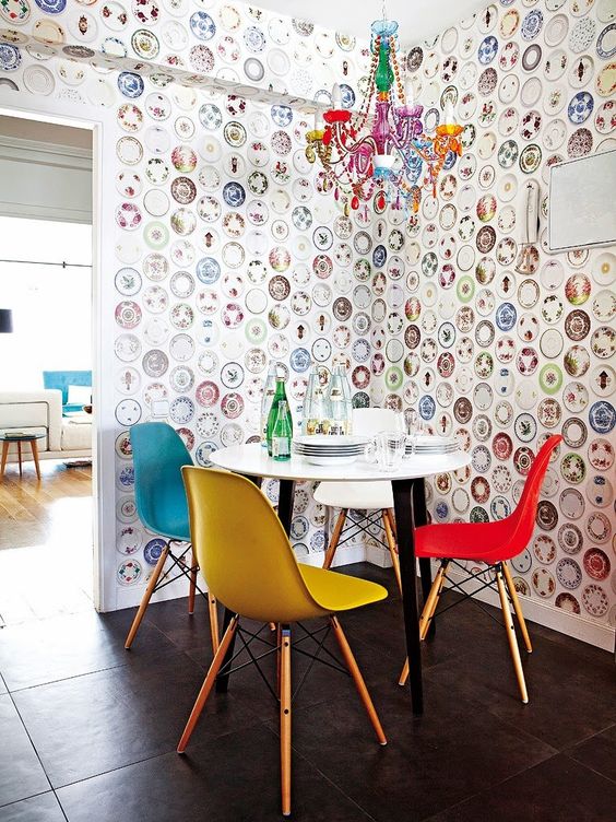 a colorful dining nook with a round table and colorful Eames chairs, a bold chandelier and catchy wallpaper with bright plates printed on it