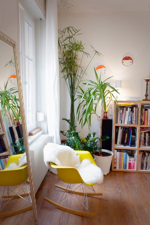 a colorful nook with a floor mirror, a bold yellow Eames rocker, a bookshelf with colorful books and potted plants is very cozy