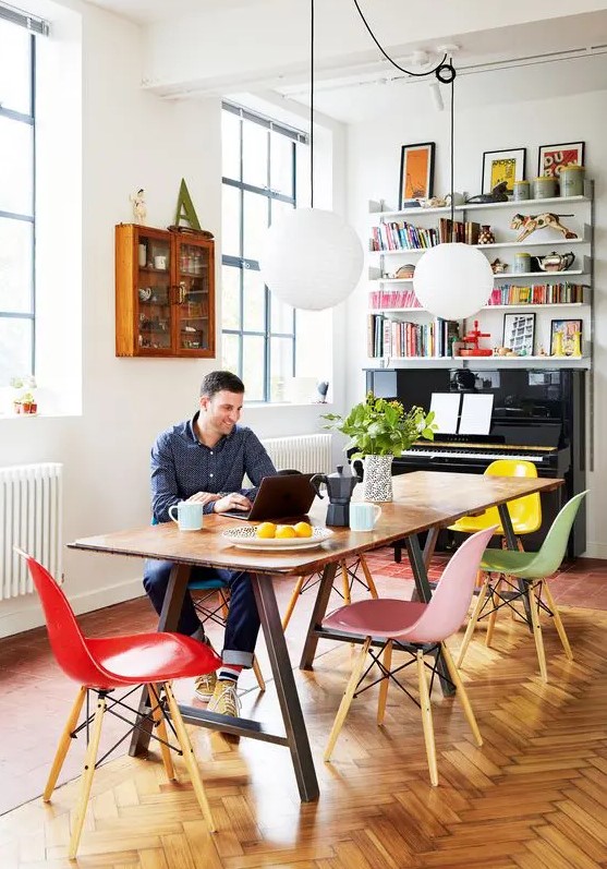 a colorful working and dining space with a trestle table, colorful Eames chairs, pendant lamps, a piano and shelves over it and a glass cabinet