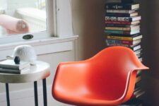a cool reading nook with a tall book stack, an orange Eames rocker, a side table with decor and a rug