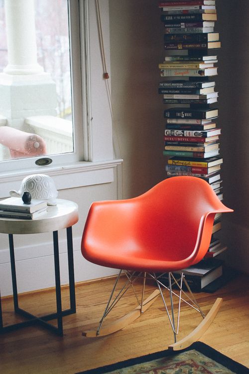 a cool reading nook with a tall book stack, an orange Eames rocker, a side table with decor and a rug