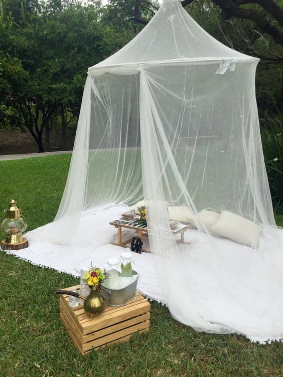a cozy outdoor space with a blanket, a couple of pillows, a little table, a mosquito net canopy, a crate with blooms