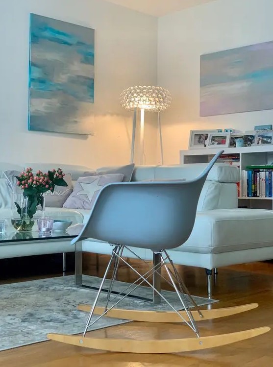 a delicate living room with a white sectional, a white Eames rocker, a glass coffee table and some pastel artwork