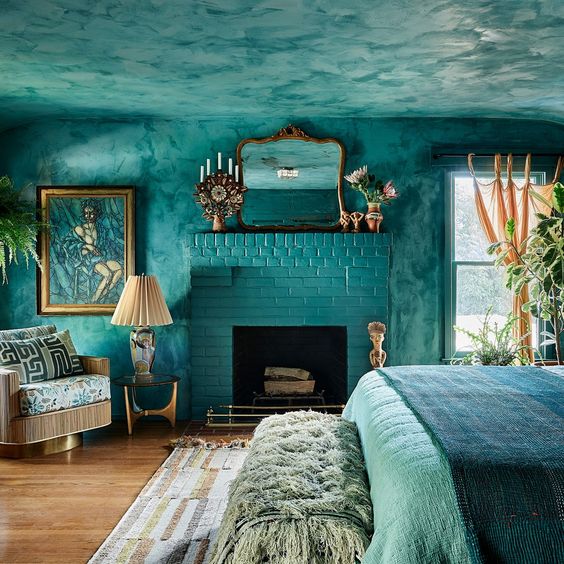 a jaw dropping turquoise bedroom with a fireplace, a bed and a bench, a printed chair and some artwork and plants