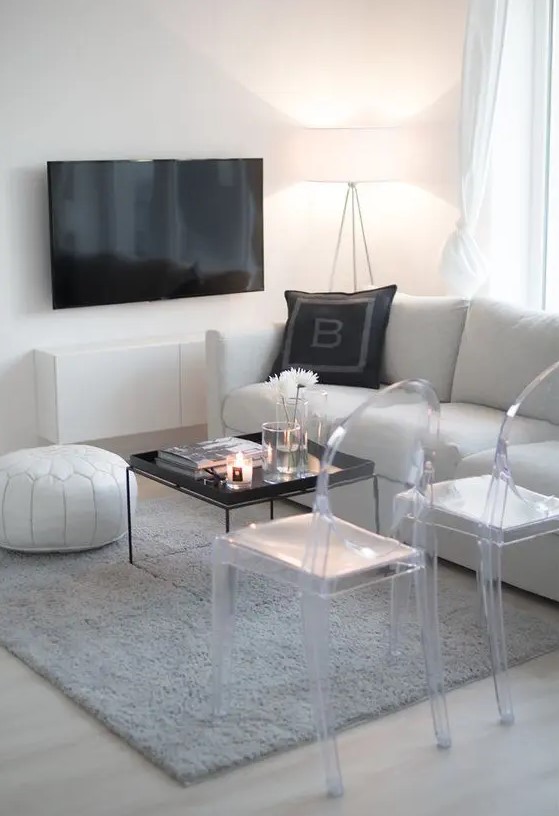 a laconic white living room with a sofa, a leather pouf, ghost chairs, a grey rug, a TV and a wall-mounted TV unit