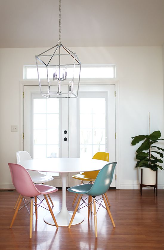 a lively dining space with a round table, colored chairs and a white Eames one, a framed pendant lamp and some potted plants