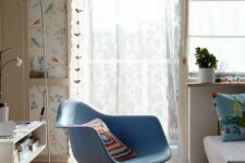a lively space with a blue Eames chair, a daybed with printed pillows, printed wallpaper, neutral curtains and a storage unit