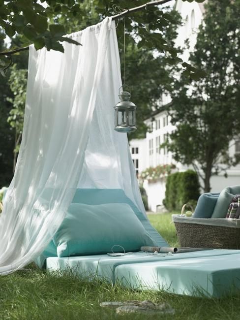a lovely outdoor space with a daybed placed right on the ground, a canopy of mosquito net, a candle lantern and a basket